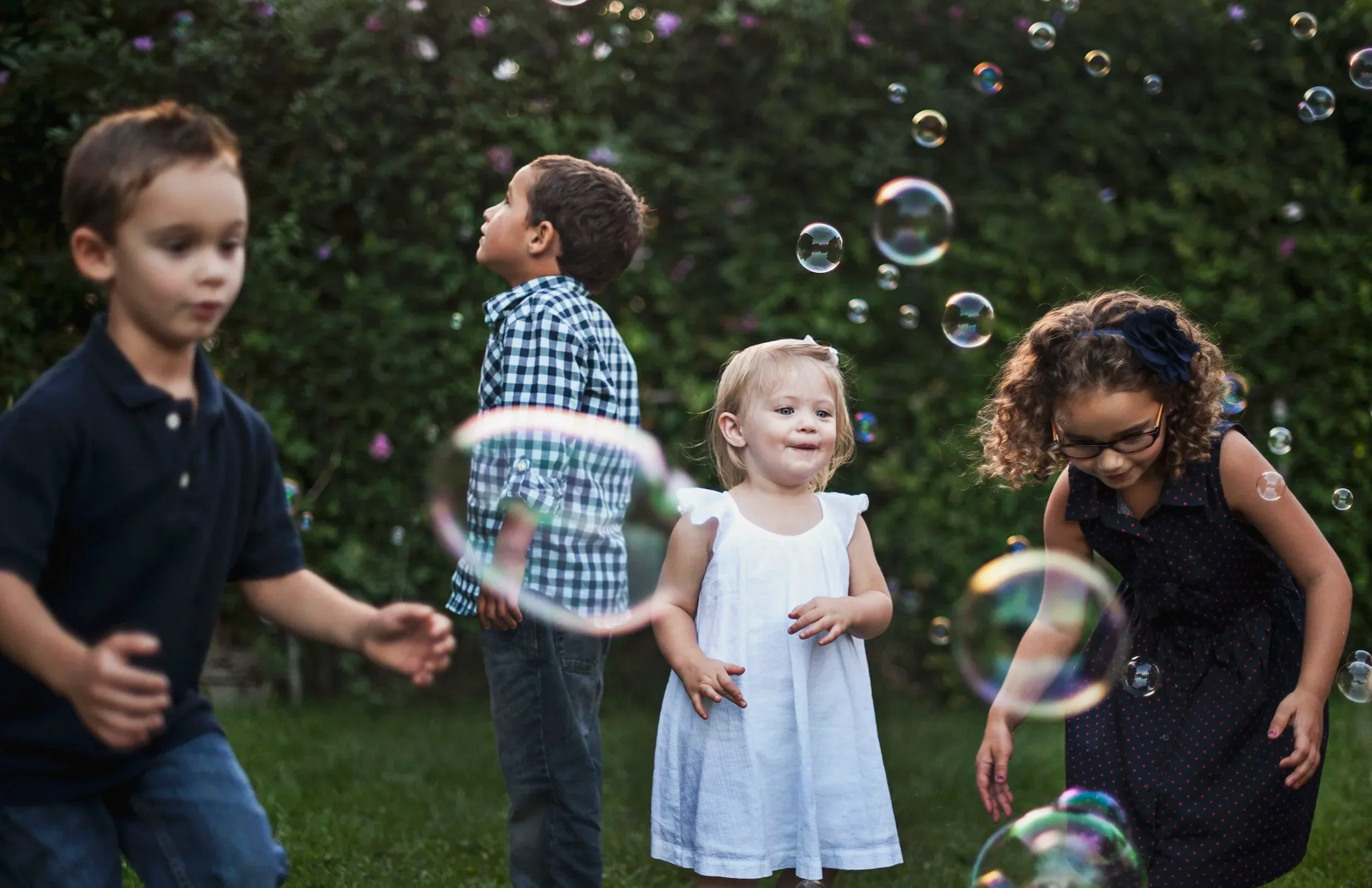 Four young children playing outdoors on the grass, chasing soap bubbles